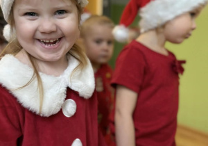 Uśmiechnięte dziecko w czerwonej świątecznej sukience i czapce Mikołaja stoi w kolejce, trzymając za rękę koleżankę z grupy.