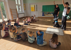 Dzieci siedzące na podłodze zgłaszają się do instruktorki pokazującej planszę obok dużej czarnej maskotki psa.