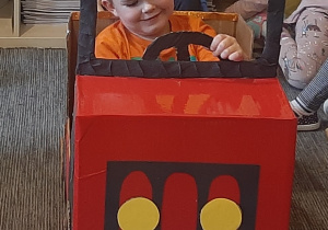 Kacper siedzi za kierownicą czerwonego auta z kartonu.