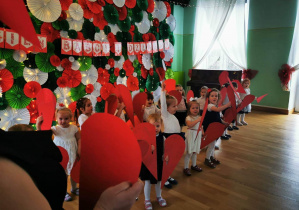 Dzieci stoją na sali gimnastycznej, trzymają w rączkach tekturowe serca -prezentują taniec.