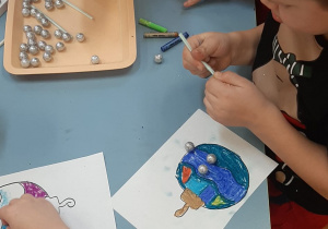 Dzieci wykonują zadanie "B jak bombka" - za pomocą słomek przenoszą małe styropianowe bombki na swoje prace i przyklejają je.