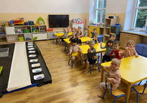 Zdjęcie przedstawiające dzieci siedzące przy stolikach przed rozpoczęciem zajęć sensorycznych.