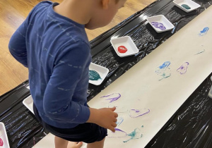 Zdjęcie przedstawiające chłopca podczas malowania stopami na papierze rozłożonym na podłodze.