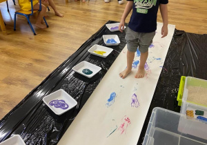 Zdjęcie przedstawiające chłopca podczas malowania stopami na papierze rozłożonym na podłodze.