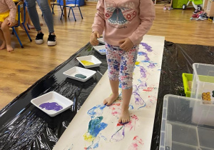 Zdjęcie przedstawiające dziewczynkę podczas malowania stopami na papierze rozłożonym na podłodze.