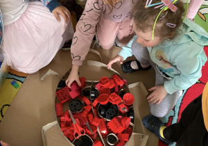 Dzieci w przedszkolnej sali wypełniają szablon biedronki, przedmiotami w kolorze czerwonym i czarnym ( klockami, zabawkami)