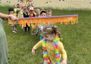 Zdjęcie przedstawiające dzieci w ogrodzie przedszkolnym podczas przechodzenia pod tyczką "Taniec Limbo".