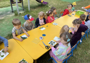 Zdjęcie przedstawia grupę dzieci siedzących przy stolikach w ogrodzie przedszkolnym.