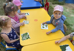 Zdjęcie przedstawia grupę dzieci siedzących przy stolikach w ogrodzie przedszkolnym.