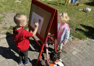 Zdjęcie przedstawiające dzieci malujące obrazy na sztalugach w ogrodzie przedszkolnym.