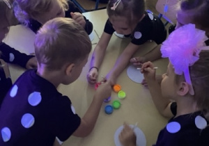 Dzieci siedzą przy stoliku i maczają patyczki kosmetyczne w fluorescencyjnych farbach, następnie malują oklejone białym papierem płyty CD.