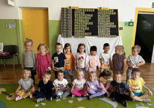 Zdjęcie przedstawiające całą grupę na holu przedszkolnym na tle tablicy odlotów i przylotów.