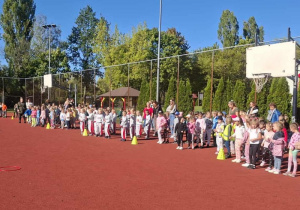Kilka grup dzieci ustawionych w rzędach na boisku sportowym, gotowych do rozpoczęcia konkurencji.