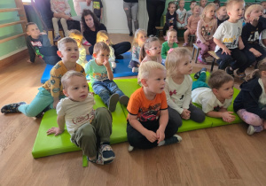 Grupa dzieci siedzi na matach i ławkach, z dużym zainteresowaniem oglądając przedstawienie.