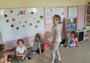 Dziewczynka przy tablicy z kolorowymi kropkami, reszta dzieci siedzi na dywanie i przygląda się zabawie.