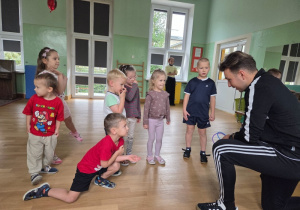 Grupa przedszkolaków rozmawia z trenerem, który kuca na wysokości ich wzroku i pokazuje im przyrząd sportowy.
