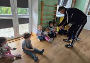 Dzieci siedzą w rzędzie na podłodze, a trener rozdaje im kolorowe woreczki gimnastyczne.