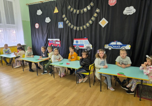 Dzieci siedzą przy stolikach na sali gimnastycznej i słuchają zasad zabawy.