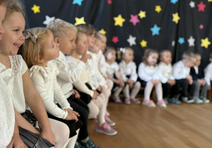 Dzieci w strojach odświętnych siedzą w rzędzie przed uroczystością.