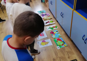 Dzieci oglądają prace konkursowe „Owocowy superbohater” ułożone na podłodze.