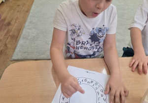 Dziecko siedzi przy stole i wskazuje na kartę pracy z literami ułożonymi wokół dużego owalu.