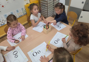 Sześć dzieci siedzi przy stole, każde z kartą pracy z literą „O” i rysunkiem owieczki.