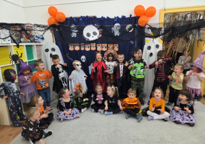 Duża grupa przedszkolaków w halloweenowych strojach pozuje przed dekoracją z napisem „HALLOWEEN”.