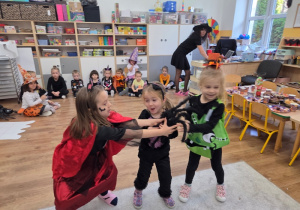 Trzy dziewczynki w przebraniach (czarownica, kot, zombie) bawią się pluszowym, dużym pająkiem; reszta dzieci siedzi i kibicuje.