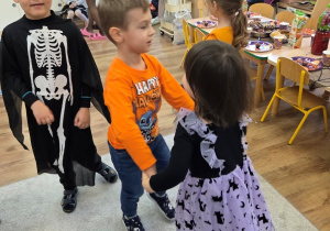 Chłopiec w koszulce „Happy Halloween” tańczy w parze z dziewczynką w fioletowej sukience w kotki; obok stoi chłopiec w stroju szkieletu.