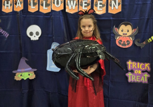 Dziewczynka przebrana za wampirzyce pozuje solo na tle dekoracji, trzyma wielkiego nadmuchiwanego pająka.