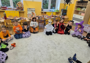 Grupa dzieci siedzi w kole na dywanie, pokazując straszne miny z pozami; za nimi stół z przekąskami.