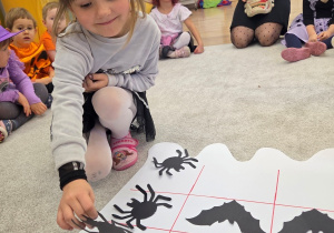 Dziewczynka układa „kółko-krzyżyk” z papierowymi pająkami i nietoperzami na dużej planszy.