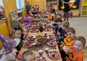 Długi stół pełen halloweenowych talerzyków i napojów; dzieci w przebraniach machają do aparatu podczas poczęstunku.