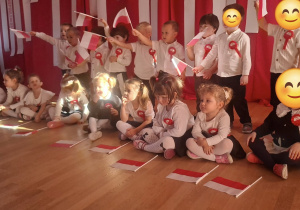 Grupa dzieci, stojąca i siedząca, macha wesoło w górze małymi polskimi flagami.