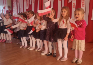 Dzieci w rzędzie, ubrane w biało-czarne stroje z dodatkami czerwieni, radośnie machają małymi polskimi flagami.