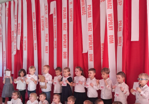 Dwie grupy dzieci stojące i siedzące. Wszystkie ubrane w białe bluzki, trzymają w rękach dyplomy.