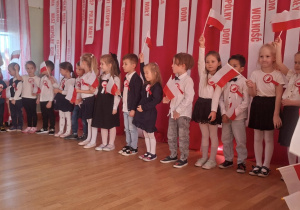 Dzieci stoją w rzędzie i podnoszą małe polskie flagi podczas występu.