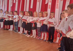 Duży rząd odświętnie ubranych dzieci. Każde dziecko trzyma na wysokości klatki piersiowej małe, biało-czerwone serce.