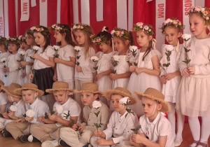 Dziewczynki w białych sukienkach i wiankach oraz chłopcy w słomkowych kapeluszach i jasnych strojach, trzymają białe róże.