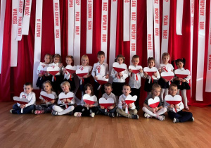 Urocze zdjęcie dwóch rzędów dzieci - siedzącego z przodu i stojącego z tyłu – które wspólnie trzymają duże, papierowe serca w barwach polskiej flagi.