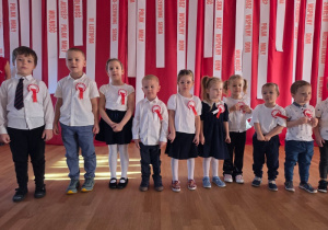 Dziewięcioro dzieci, odświętnie ubranych w białe koszule i eleganckie dodatki (krawaty, spódniczki, kokardy), stoi w rzędzie, śpiewając piosenkę patriotyczną.