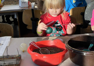 Dziecko siedzi przy stole i maluje bombkę zieloną farbą, wokół leżą materiały plastyczne.