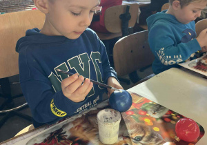 Chłopiec w niebieskiej bluzie ozdabia niebieską bombkę, pracując przy stole w jasnej sali.