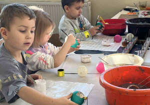 Kilkoro dzieci siedzi przy stole i dekoruje kolorowe bombki różnymi farbami i brokatem.