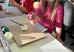 Dziewczynka uśmiecha się, trzymając w ręku czerwoną bombkę pokrytą brokatem; obok inne dzieci pracują nad ozdobami.