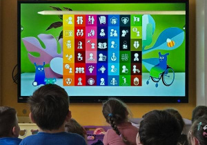 Dzieci siedzą przed dużym ekranem, na którym wyświetlane są kolorowe ikony przedstawiające prawa dziecka.