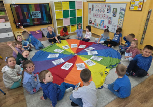 Grupa dzieci siedzi wokół kolorowej chusty animacyjnej, na której leżą obrazki związane z prawami dziecka.