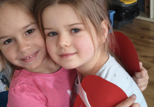 Dwie dziewczynki przytulają się i uśmiechają, trzymając duże czerwone serce z papieru.