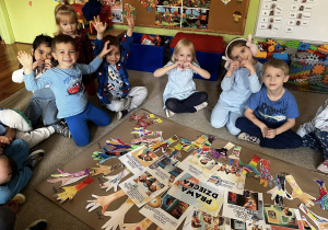 Kilka dzieci siedzi na dywanie wokół dużej planszy z rysunkami dłoni i obrazkami przedstawiającymi prawa dziecka.