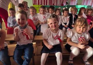 Grupa dzieci siedzi na ławkach, ubrane odświętnie w białe bluzki i koszule z biało-czerwonymi kotylionami.